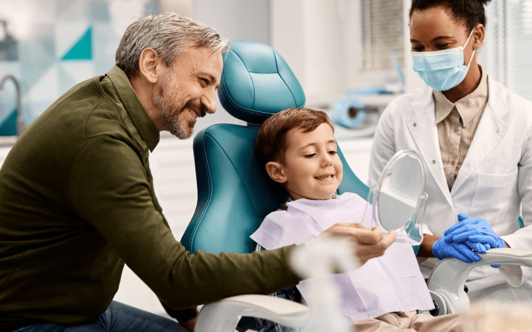 Young boy sitting in chair at the dentist, father is smiling on the left, holding a mirror for son to look at teeth, dentist on right smiling at boy.
